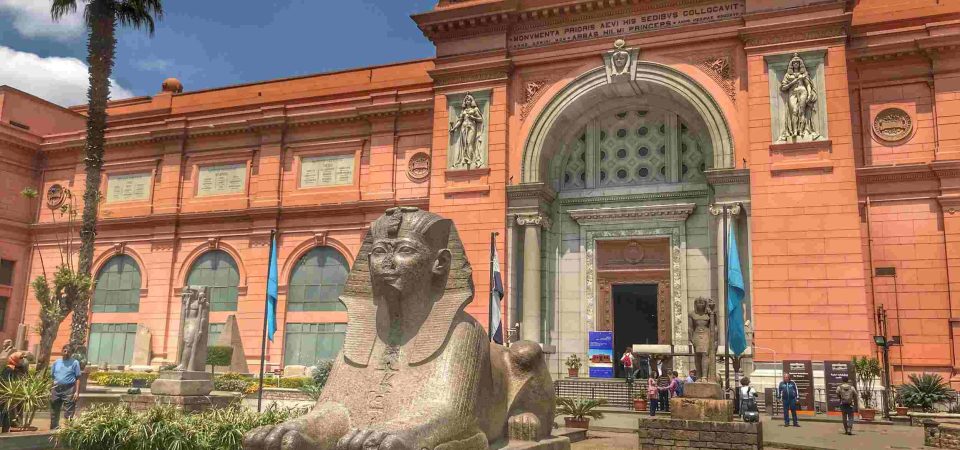 Front view of the Egyptian Museum in Cairo with a large ancient sphinx statue in the courtyard under a partly cloudy sky