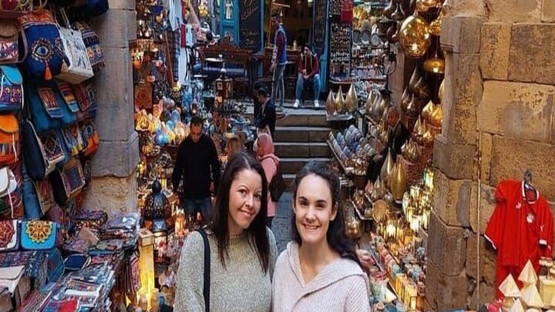 Two smiling women standing in a vibrant alley of Khan El Khalili market in Cairo, surrounded by colorful bags, lamps, and Egyptian souvenirs